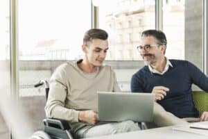 Mature businessman and young man in wheelchair using laptop in office