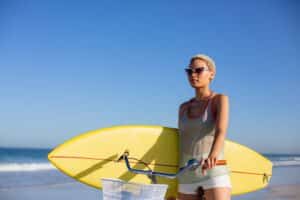 Front view of African american woman with surfboard sitting on bicycle at beach in the sunshine